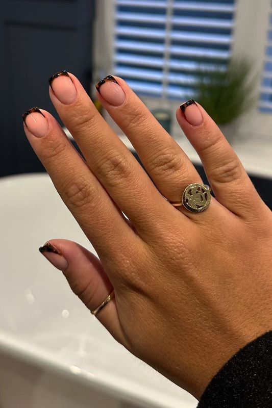 Nägel mit nudefarbener Basis und schwarzen Spitzen, getragen mit zwei Ringen; ein Ring mit rundem Design und ein feines Band.